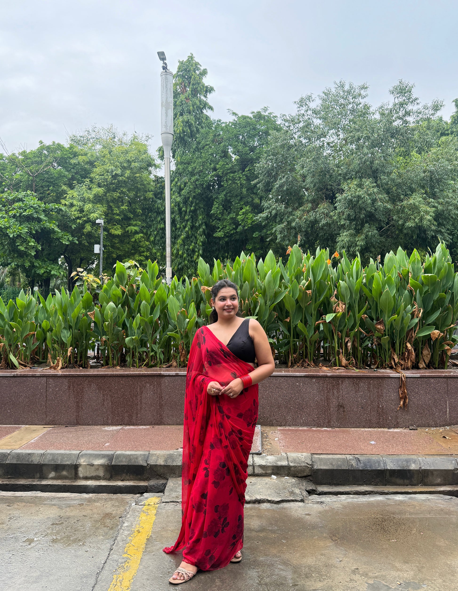 Woman in a red saree standing outdoors with greenery in the background