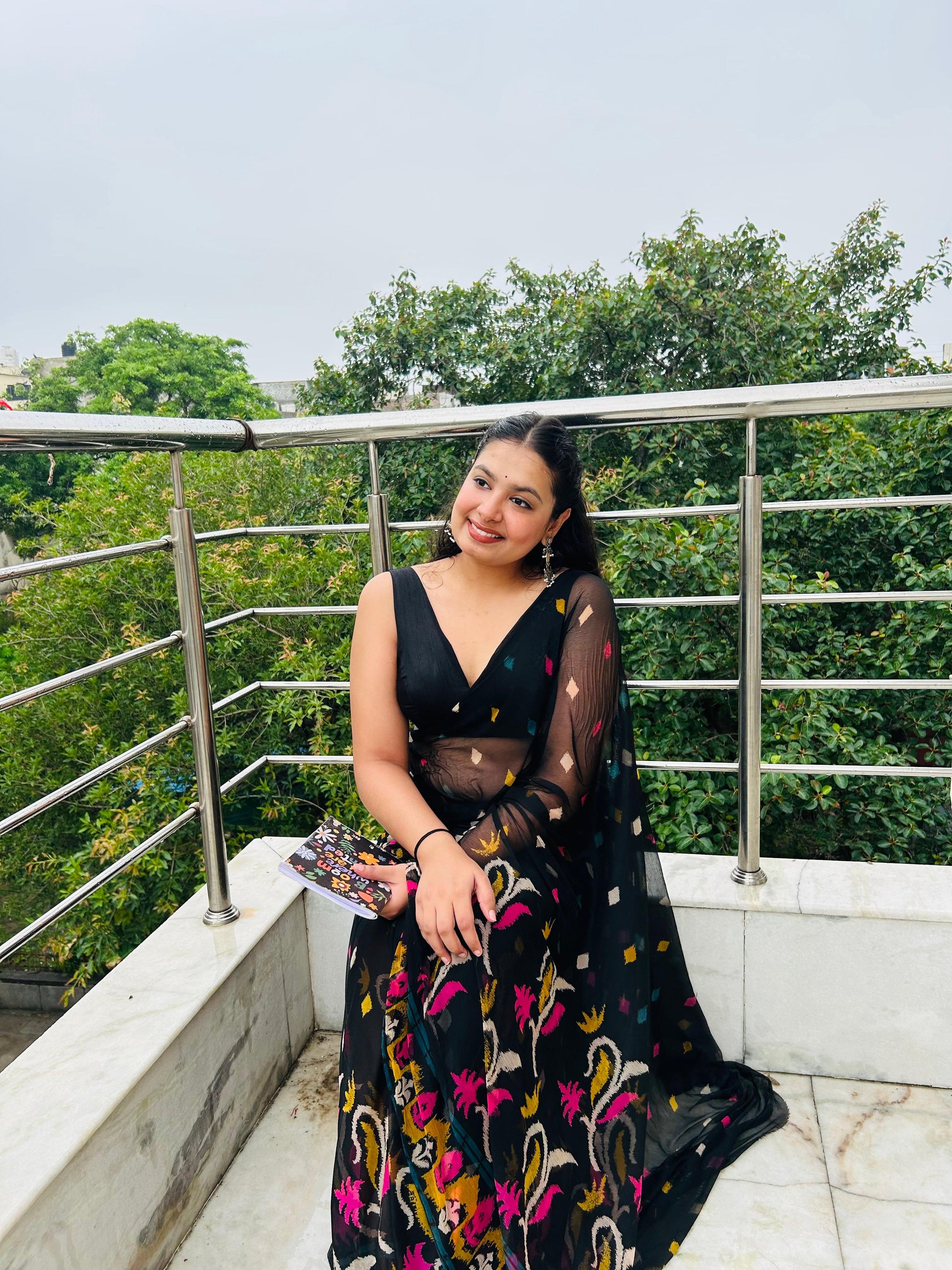 Woman in a black floral dress standing on a balcony with green trees in the background