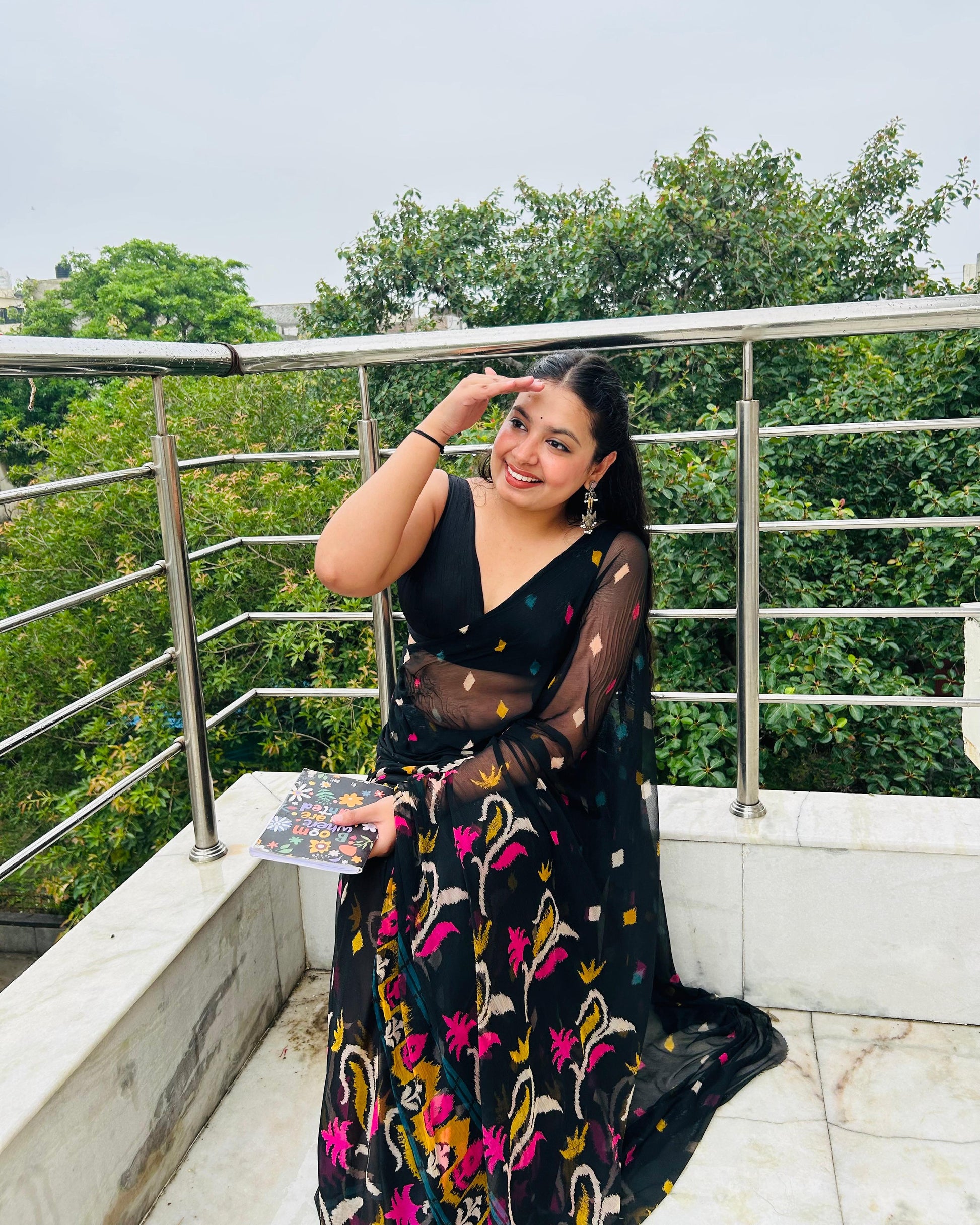 Woman in a black floral saree on a balcony with green trees in the background