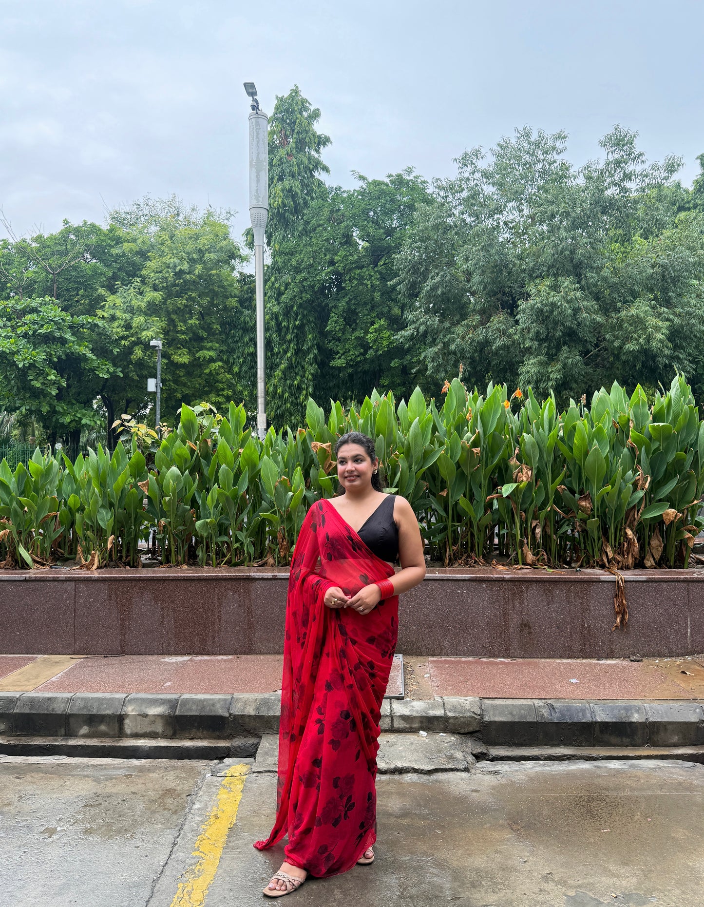 Woman in a red saree standing outdoors with greenery in the background