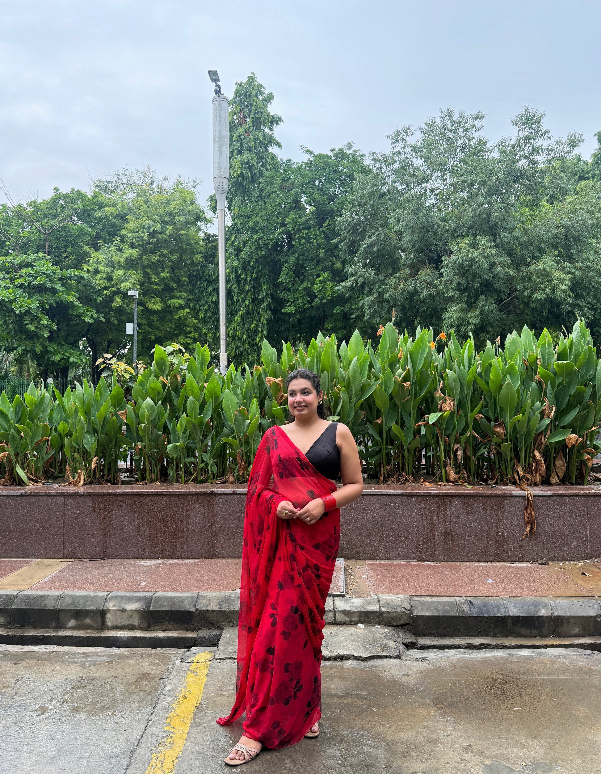 Woman in a red saree standing outdoors with greenery in the background
