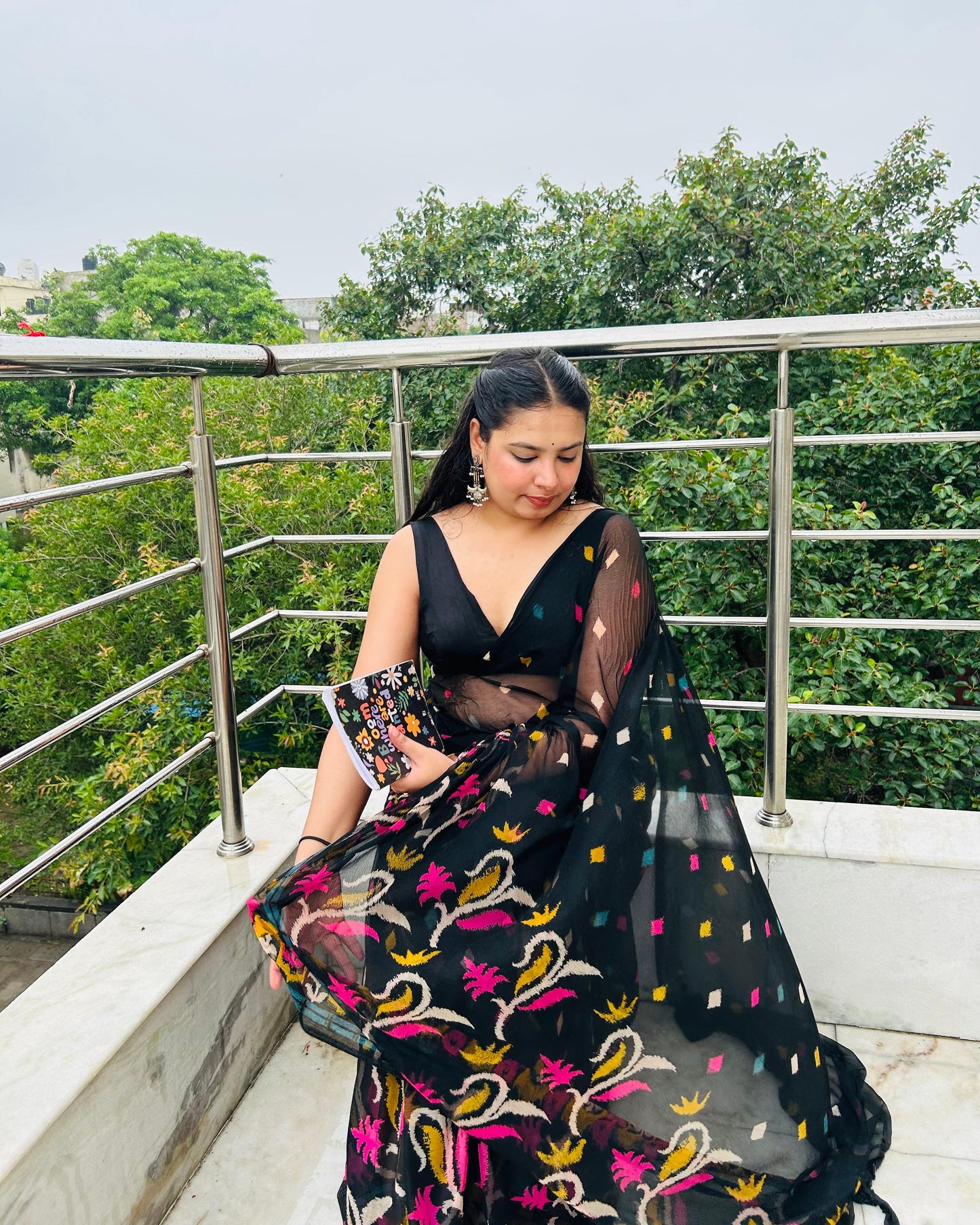 Woman in a black and colorful traditional outfit on a balcony with greenery in the background