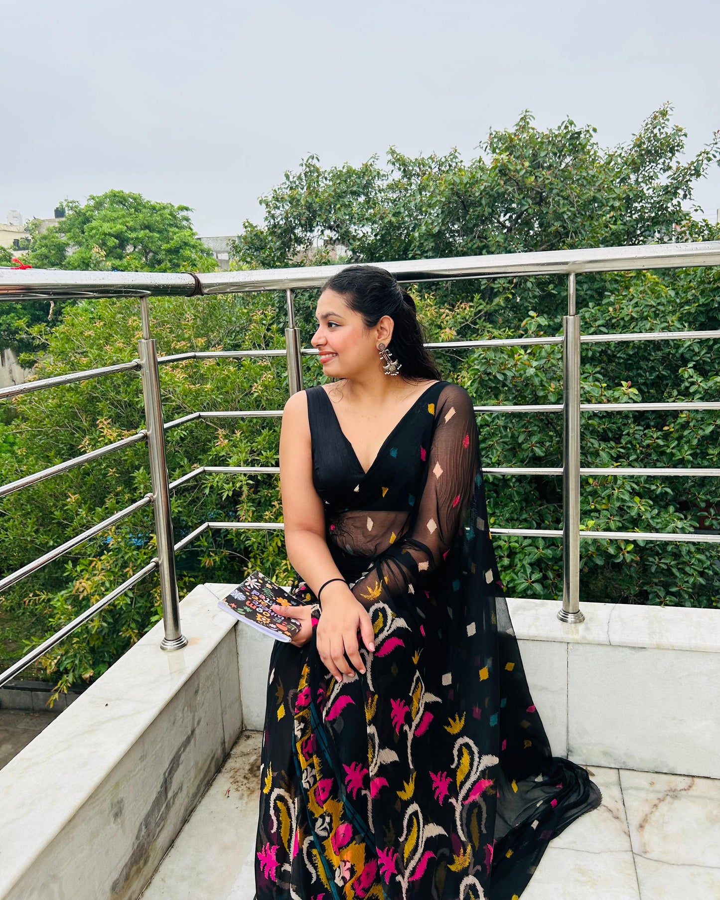 Woman in a black floral dress sitting on a balcony with green trees in the background
